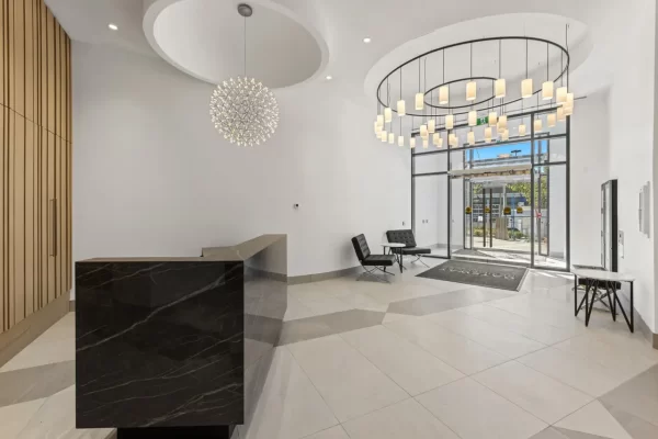 Modern luxury apartment lobby at Gateway Residences in West Vancouver, featuring a sleek dark marble reception desk, contemporary lounge seating with Barcelona-style chairs, a grand tiered contemporary chandelier, and large glass entry doors.